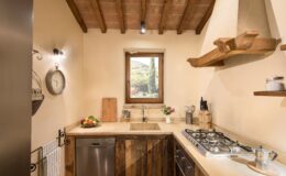 Cozy kitchen interior featuring wooden beams, a window with a scenic view, a stainless steel dishwasher, a gas stove, and decorative elements like a clock and fresh flowers.