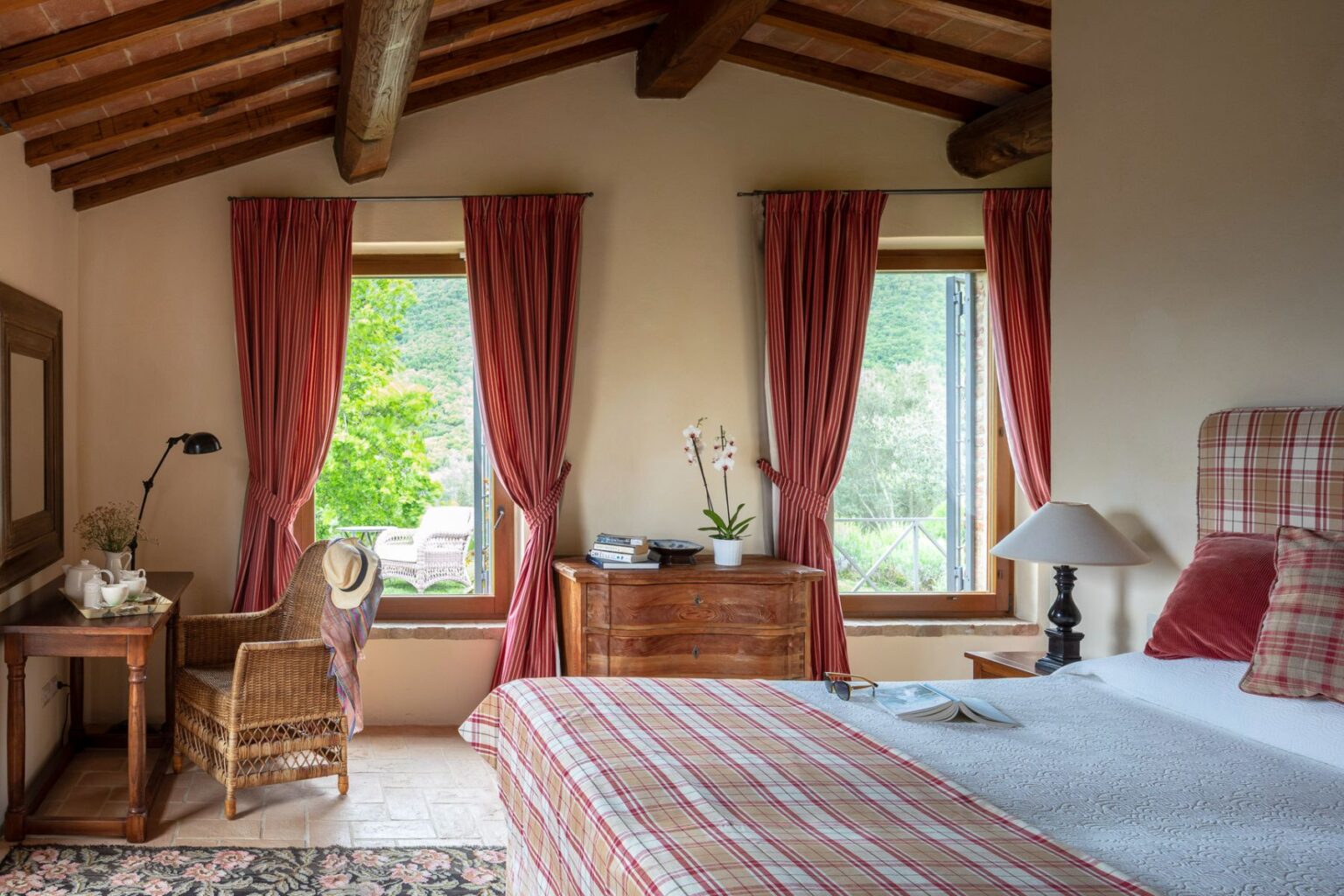 Cozy bedroom featuring rustic decor, wooden beams, and large windows with red curtains. A comfortable bed with plaid bedding, a wooden nightstand with a lamp, and a wicker chair beside a table set with tea. A view of greenery outside adds a serene touch to the inviting atmosphere. | Only on SopranoVillas