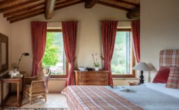 Cozy bedroom featuring rustic decor, wooden beams, and large windows with red curtains. A comfortable bed with plaid bedding, a wooden nightstand with a lamp, and a wicker chair beside a table set with tea. A view of greenery outside adds a serene touch to the inviting atmosphere.