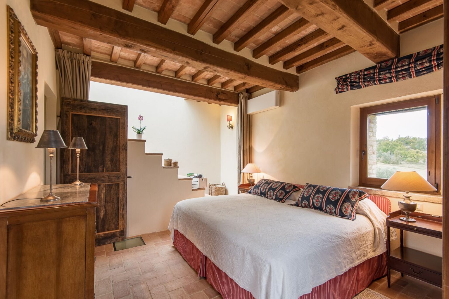 Cozy bedroom featuring a king-sized bed with decorative pillows, wooden beams on the ceiling, a rustic wooden door, and warm lighting from bedside lamps. The room includes a staircase leading to another level, a window showcasing a scenic view, and elegant furnishings that enhance the inviting atmosphere. | Only on SopranoVillas