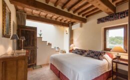 Cozy bedroom featuring a king-sized bed with decorative pillows, wooden beams on the ceiling, a rustic wooden door, and warm lighting from bedside lamps. The room includes a staircase leading to another level, a window showcasing a scenic view, and elegant furnishings that enhance the inviting atmosphere.