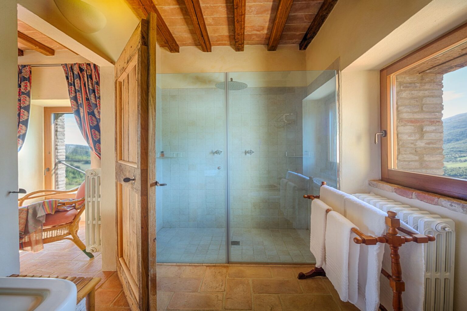 Spacious bathroom featuring a modern glass shower, rustic wooden door, and scenic views through a large window. The design includes terracotta flooring, a towel rack with white towels, and a cozy seating area with a wicker chair and colorful drapes. | Only on SopranoVillas