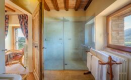 Spacious bathroom featuring a modern glass shower, rustic wooden door, and scenic views through a large window. The design includes terracotta flooring, a towel rack with white towels, and a cozy seating area with a wicker chair and colorful drapes.