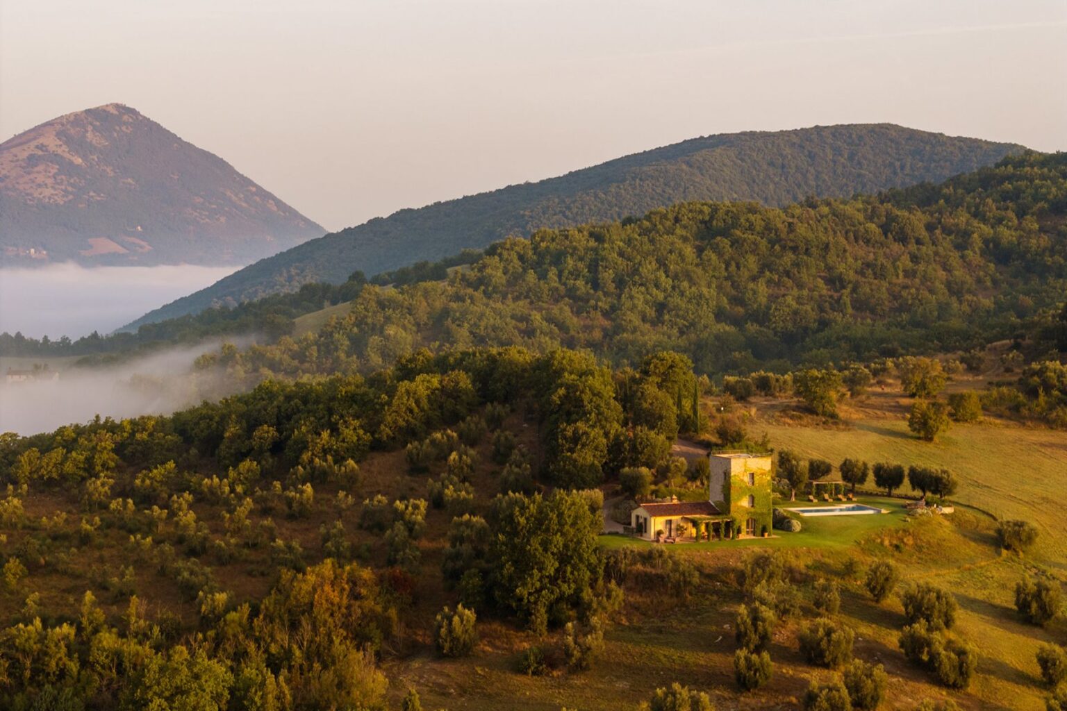 Aerial view of a rustic villa surrounded by lush green hills and mountains, with a swimming pool and olive trees in the foreground, showcasing the serene beauty of the Italian countryside at dawn. | Only on SopranoVillas
