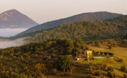 Aerial view of a rustic villa surrounded by lush green hills and mountains, with a swimming pool and olive trees in the foreground, showcasing the serene beauty of the Italian countryside at dawn.