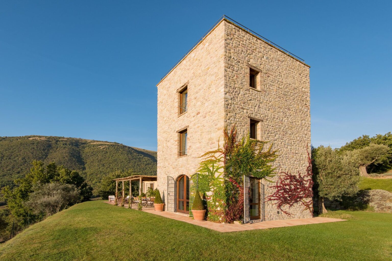 Stone house with a modern design surrounded by lush greenery and rolling hills under a clear blue sky, showcasing a spacious patio and vibrant climbing plants. Ideal for countryside retreats and architectural photography. | Only on SopranoVillas