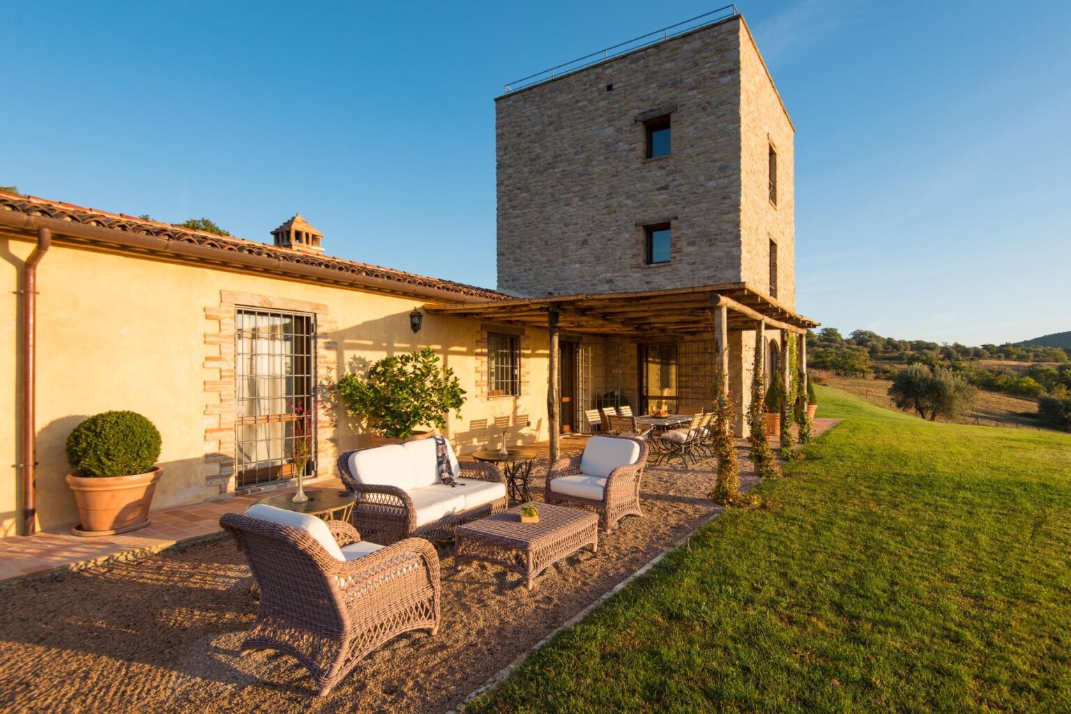 Rustic stone villa with a modern design, featuring an outdoor seating area with wicker furniture, surrounded by lush greenery and rolling hills under a clear blue sky. Ideal for relaxation and enjoying nature. | Only on SopranoVillas