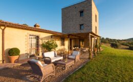 Rustic stone villa with a modern design, featuring an outdoor seating area with wicker furniture, surrounded by lush greenery and rolling hills under a clear blue sky. Ideal for relaxation and enjoying nature.