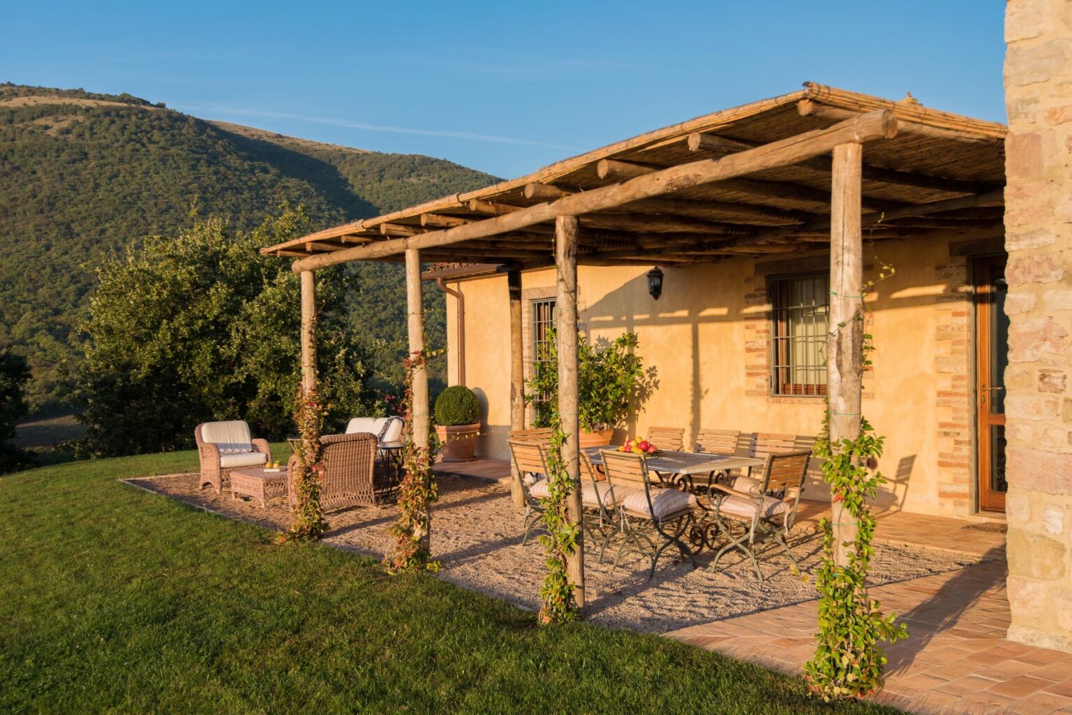 Charming outdoor patio of a rustic villa surrounded by lush green hills, featuring comfortable seating and a dining area under a wooden pergola, ideal for relaxation and enjoying nature. | Only on SopranoVillas