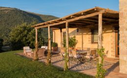 Charming outdoor patio of a rustic villa surrounded by lush green hills, featuring comfortable seating and a dining area under a wooden pergola, ideal for relaxation and enjoying nature.
