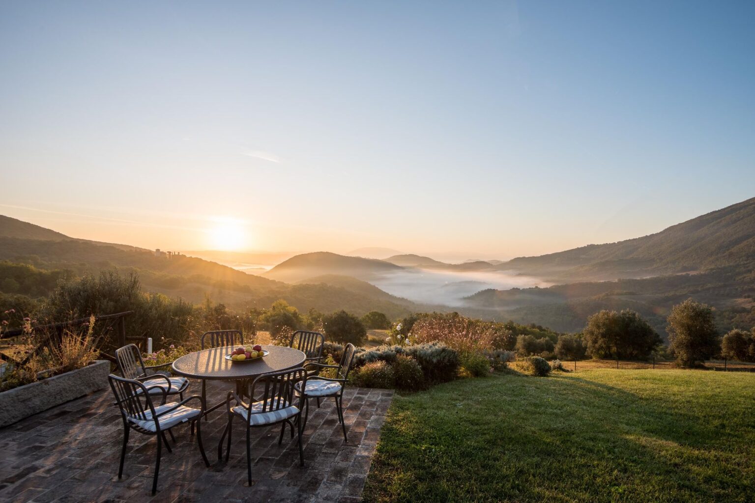 Scenic view of a sunrise over rolling hills, with a patio table set for breakfast, featuring a fruit bowl, surrounded by lush greenery and misty valleys in the background. | Only on SopranoVillas