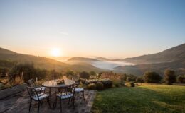Scenic view of a sunrise over rolling hills, with a patio table set for breakfast, featuring a fruit bowl, surrounded by lush greenery and misty valleys in the background.