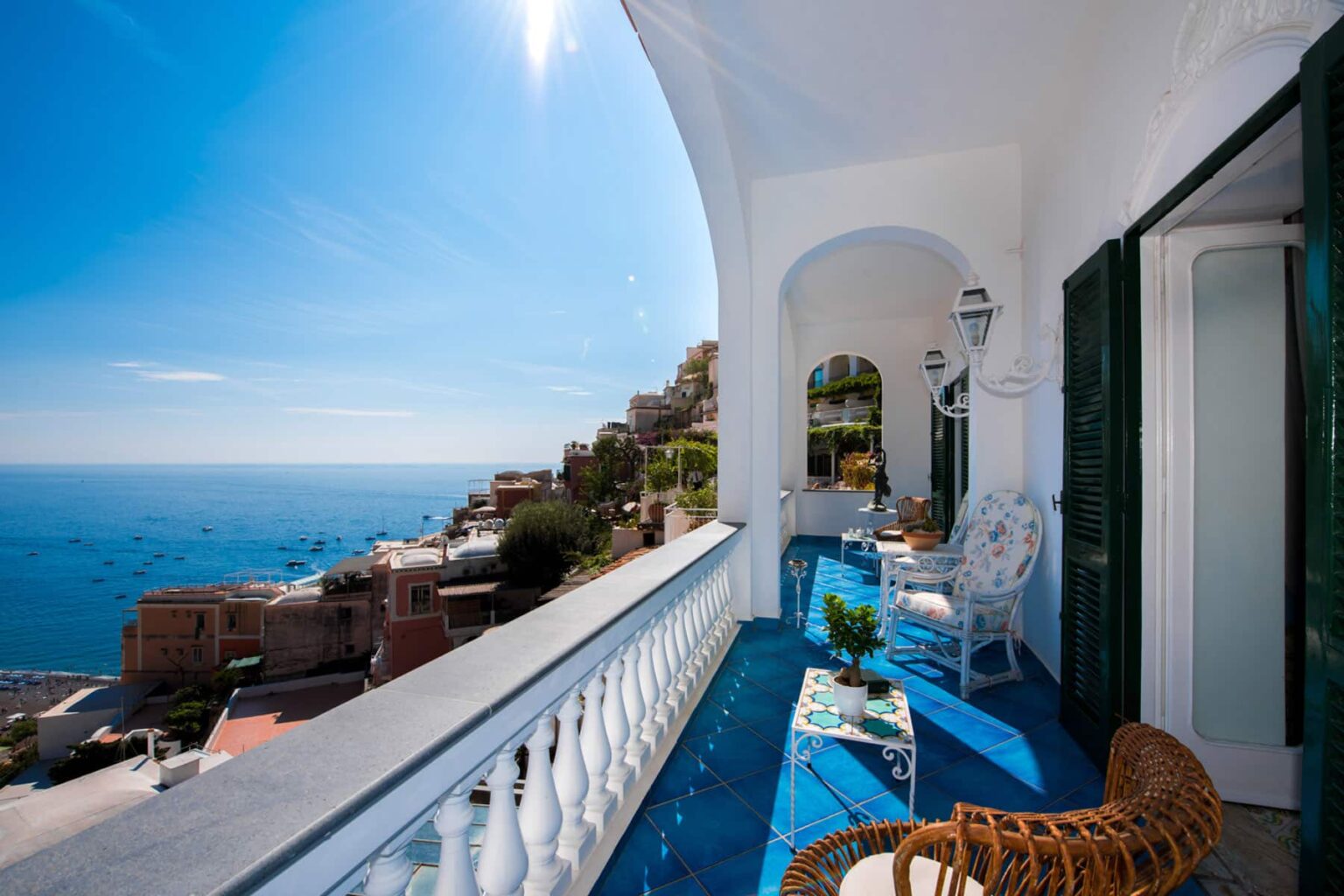 Scenic balcony view overlooking the Amalfi Coast, featuring white railing, wicker furniture, and vibrant blue tile flooring under a clear blue sky. | Only on SopranoVillas
