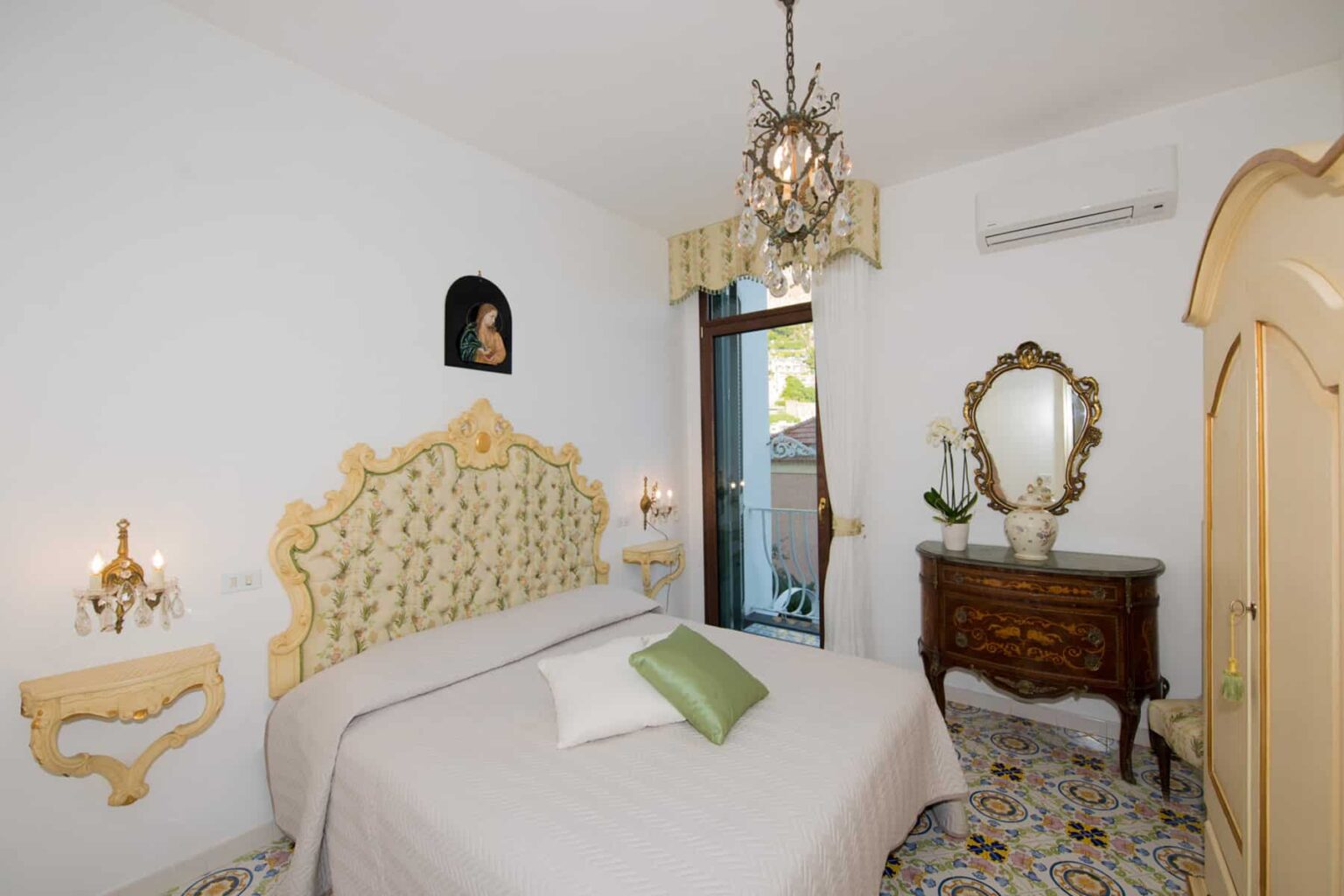 Elegant bedroom featuring a vintage-style bed with an ornate headboard, soft lighting from wall sconces, a decorative mirror, and a classic wooden dresser. The room has a bright ambiance with white walls and colorful tiled flooring, creating a cozy and inviting atmosphere. | Only on SopranoVillas