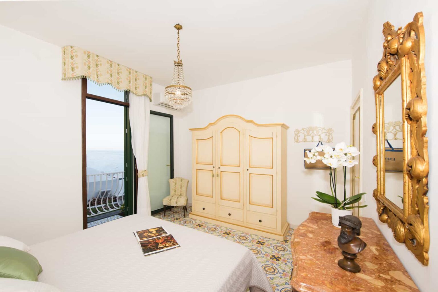 Cozy bedroom featuring an elegant vintage wardrobe, a decorative mirror, and a serene ocean view through large windows. The room is adorned with soft lighting and floral accents, creating a tranquil atmosphere. | Only on SopranoVillas
