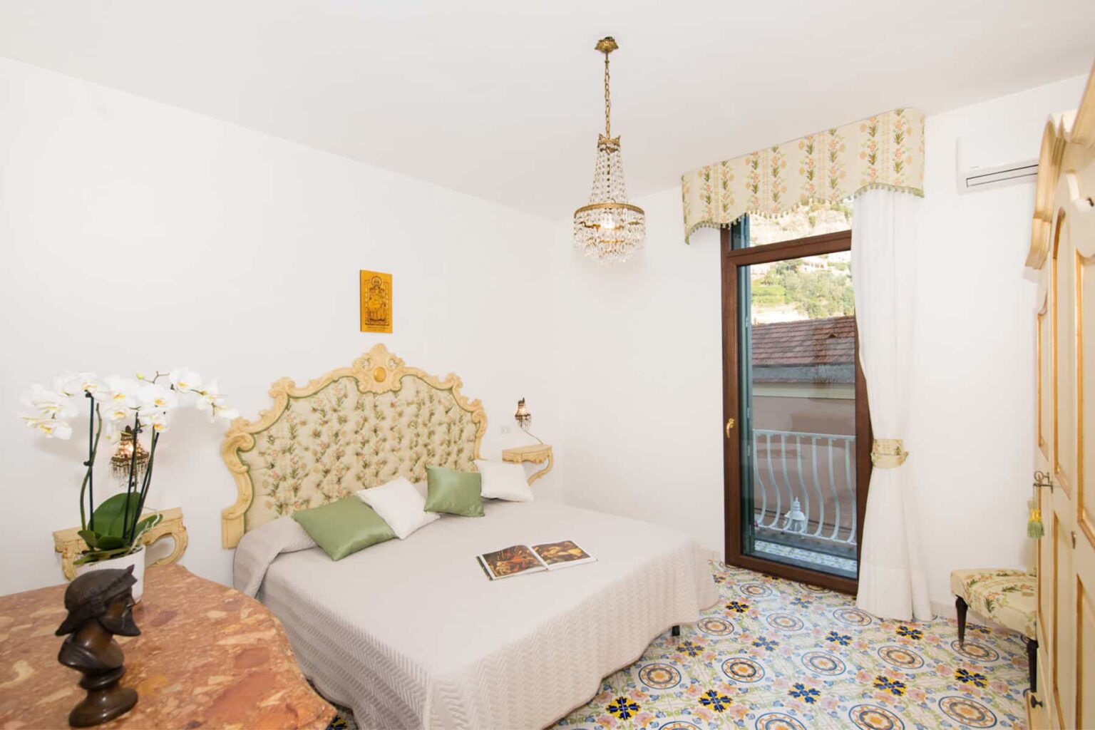 Cozy bedroom featuring a decorative bed with green accent pillows, elegant chandelier, and large window with a view. The room has a floral design on the floor and a touch of modern decor with white walls and soft lighting. Ideal for relaxation and comfort. | Only on SopranoVillas