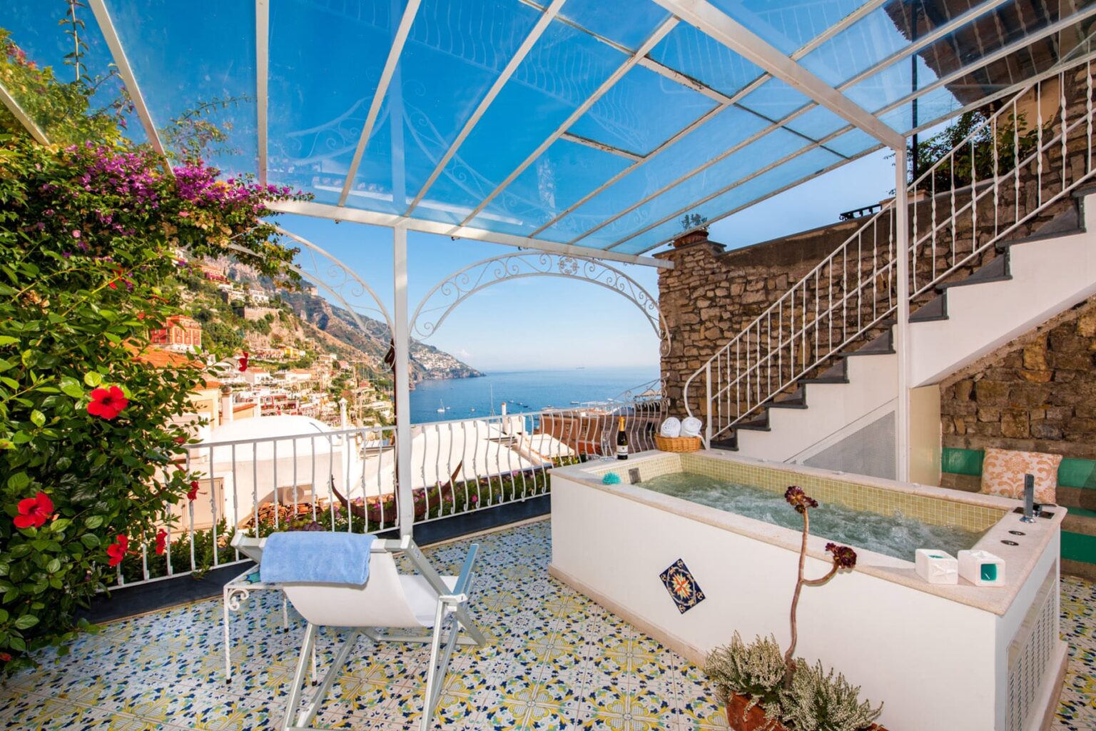 Luxurious outdoor terrace with a hot tub, surrounded by vibrant flowers and stunning sea views, featuring a clear roof and elegant tiled flooring. Ideal for relaxation and enjoying the Amalfi Coast scenery. | Only on SopranoVillas