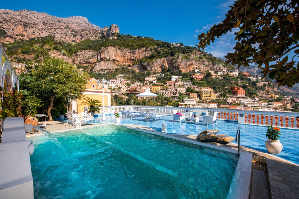 Scenic view of a luxury infinity pool overlooking a coastal village, surrounded by lush greenery and dramatic cliffs under a clear blue sky. | Only on SopranoVillas