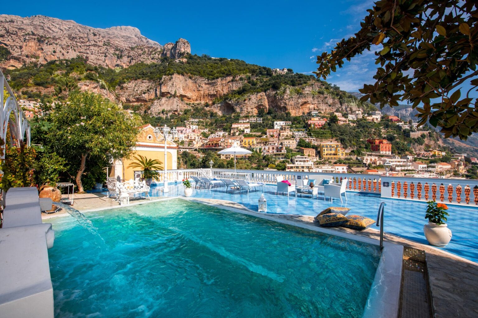 Scenic view of a luxury infinity pool overlooking a coastal village, surrounded by lush greenery and dramatic cliffs under a clear blue sky. | Only on SopranoVillas