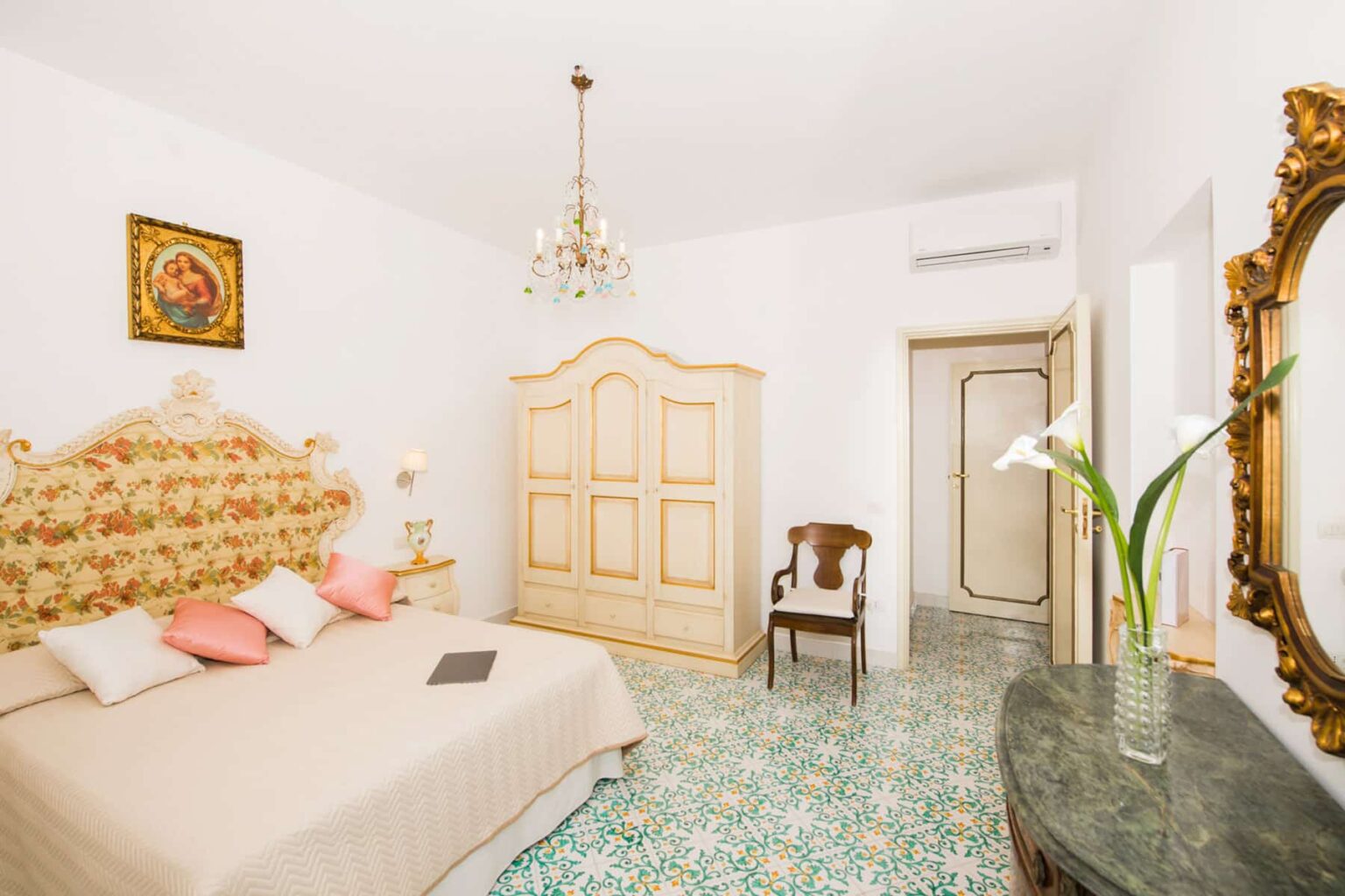 Elegant bedroom featuring a floral headboard, soft pink pillows, vintage furniture, and a decorative chandelier. The room is bright with white walls and ornate details, complemented by a patterned floor and a stylish mirror. Ideal for a cozy and sophisticated stay. | Only on SopranoVillas