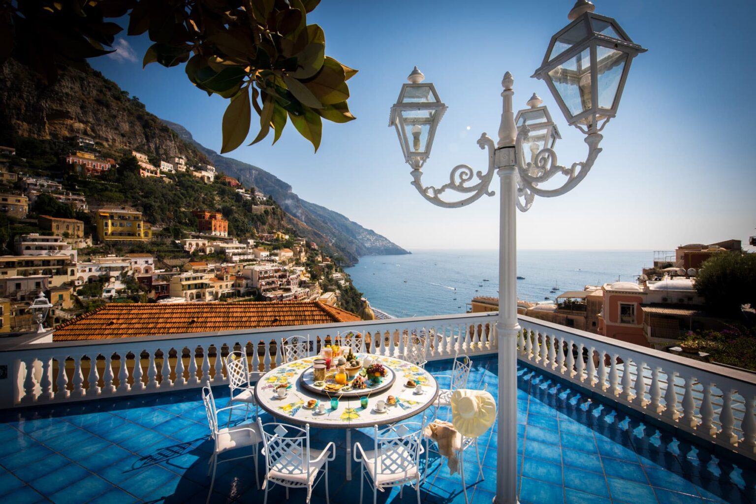 Scenic view from a terrace overlooking the Amalfi Coast, featuring a beautifully set dining table with colorful dishes and a lamp post, surrounded by lush greenery and stunning coastal cliffs. | Only on SopranoVillas