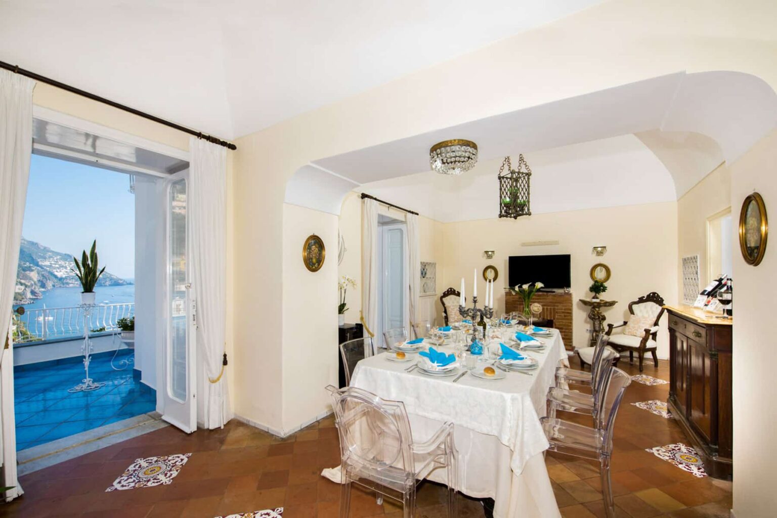 Elegant dining room with a beautifully set table featuring blue accents, surrounded by tasteful decor and large windows offering a scenic view of the sea. The light-colored walls enhance the spacious atmosphere, complemented by classic furniture and natural light. | Only on SopranoVillas