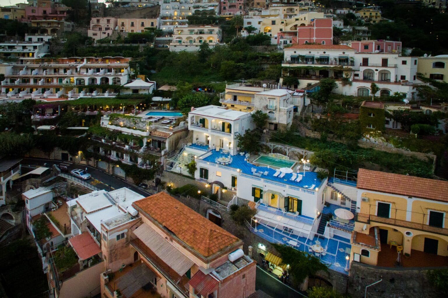 Aerial view of colorful hillside buildings in a coastal town, showcasing terraced architecture, swimming pools, and lush greenery, typical of Mediterranean landscapes. | Only on SopranoVillas