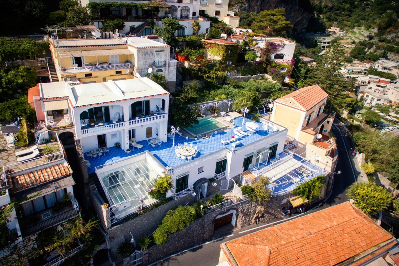 Aerial view of a luxurious villa with a blue rooftop and swimming pool, surrounded by lush greenery and residential buildings on a hillside. | Only on SopranoVillas