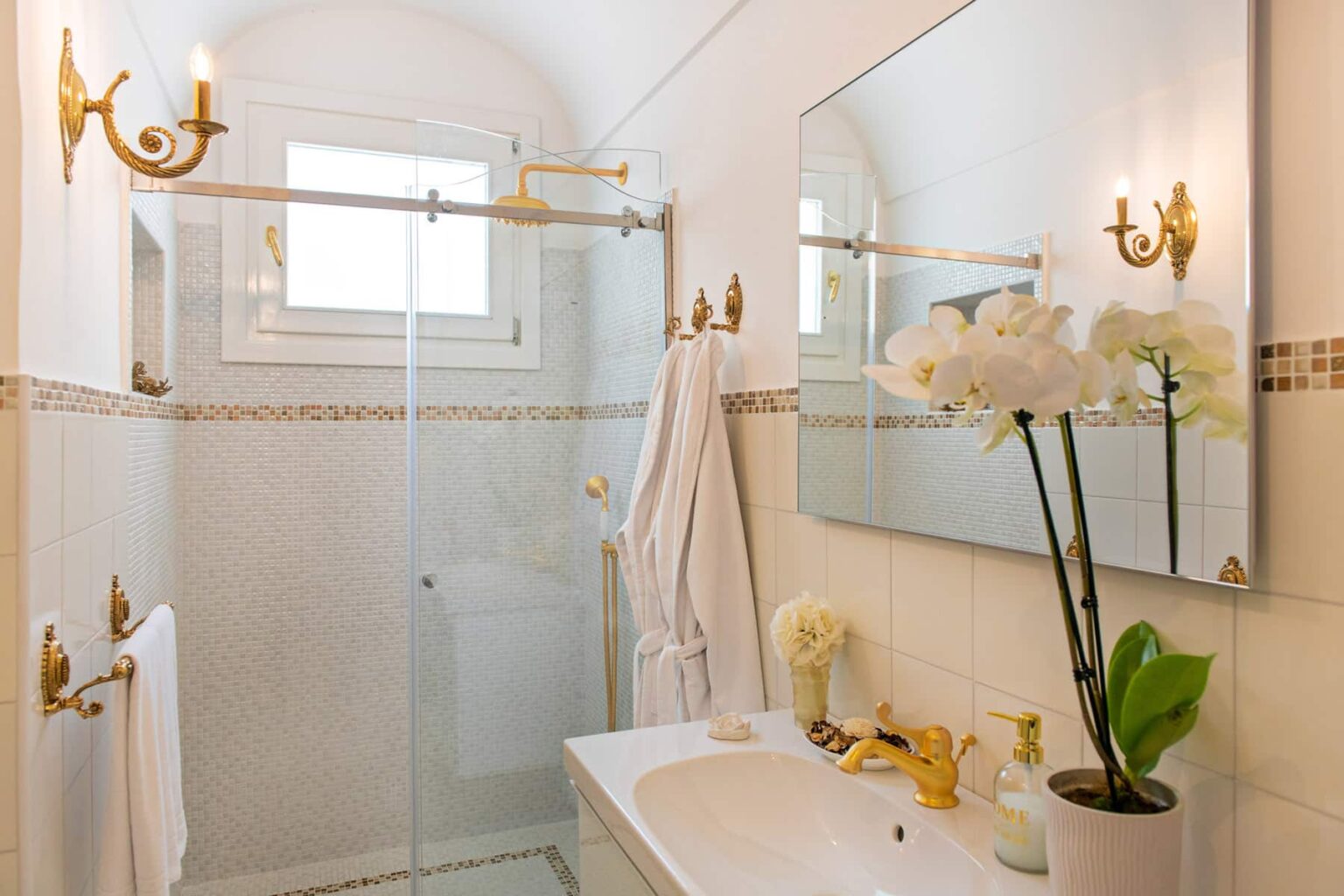 Elegant bathroom featuring a glass shower, modern fixtures, and decorative plants. The space is illuminated by stylish wall sconces and includes a large mirror, creating a bright and inviting atmosphere. | Only on SopranoVillas