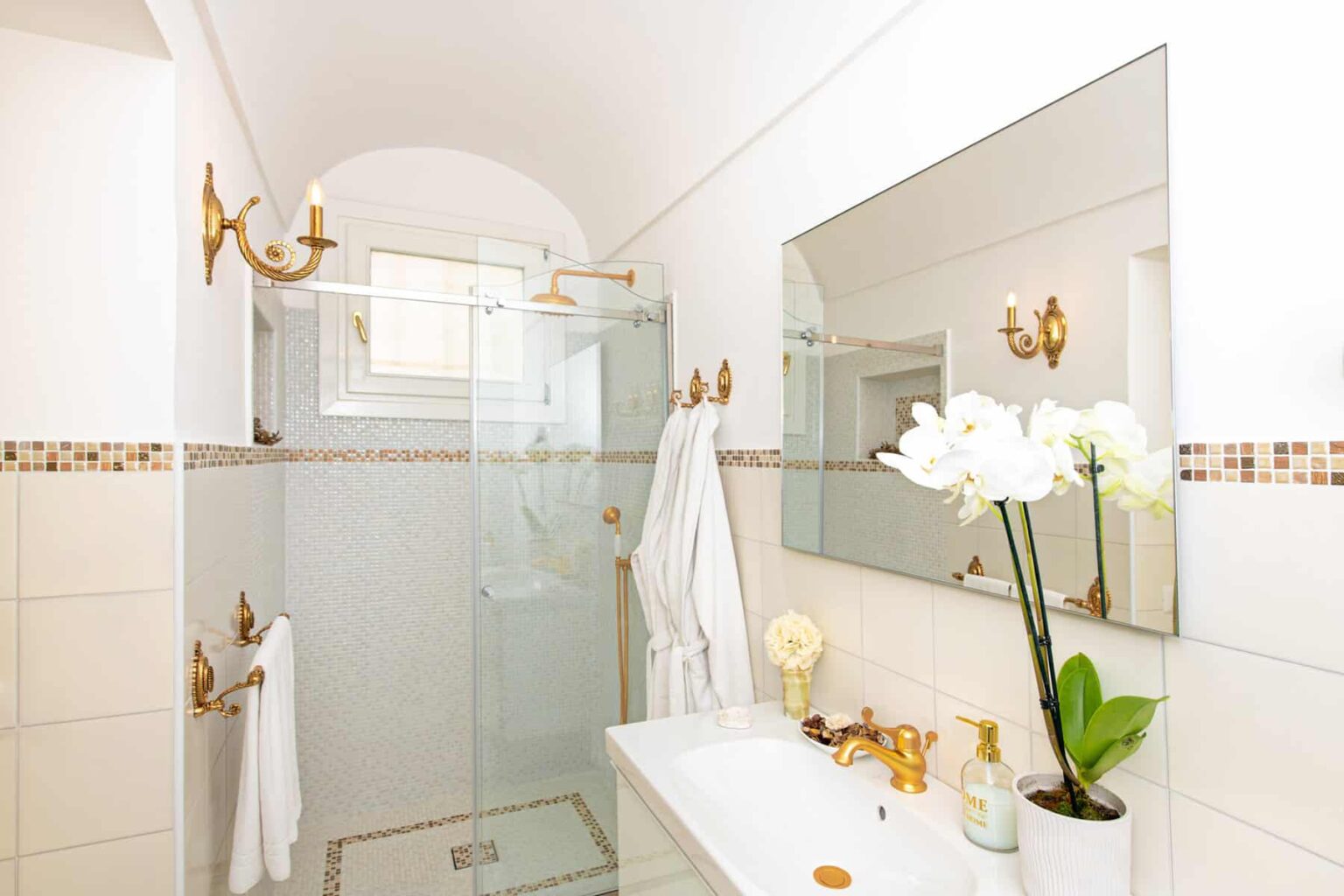 Modern bathroom featuring a glass shower, elegant gold fixtures, a large mirror, and decorative white orchids on the countertop. Bright and airy design with neutral tiles and soft lighting. | Only on SopranoVillas