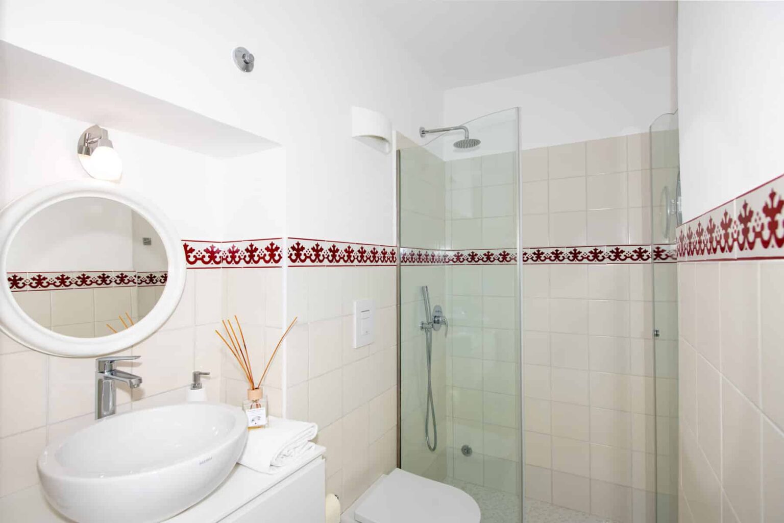 Modern bathroom featuring a glass shower enclosure, circular mirror, and elegant decorative tiles. The space is well-lit and designed for functionality and style, showcasing a clean and contemporary aesthetic. | Only on SopranoVillas