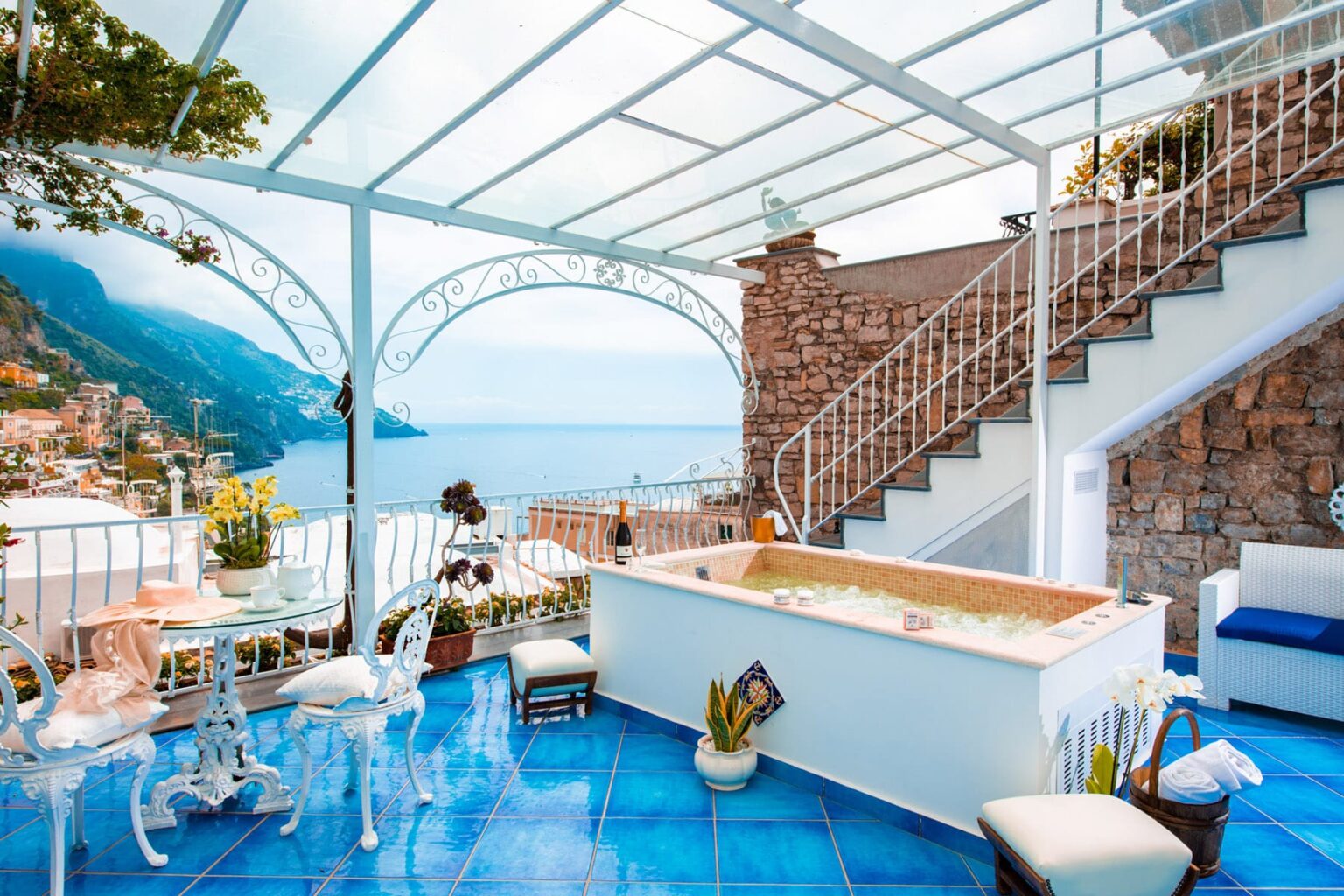 Luxurious terrace with a hot tub overlooking the Amalfi Coast, featuring blue tiled flooring, elegant white furniture, and a scenic view of the sea and mountains. | Only on SopranoVillas