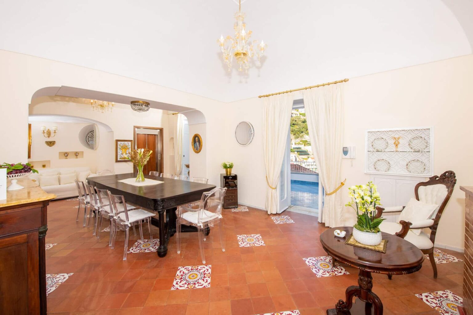 Spacious dining room featuring a large black table, transparent chairs, and elegant decor, with a view of the pool through open doors and natural light illuminating the space. | Only on SopranoVillas