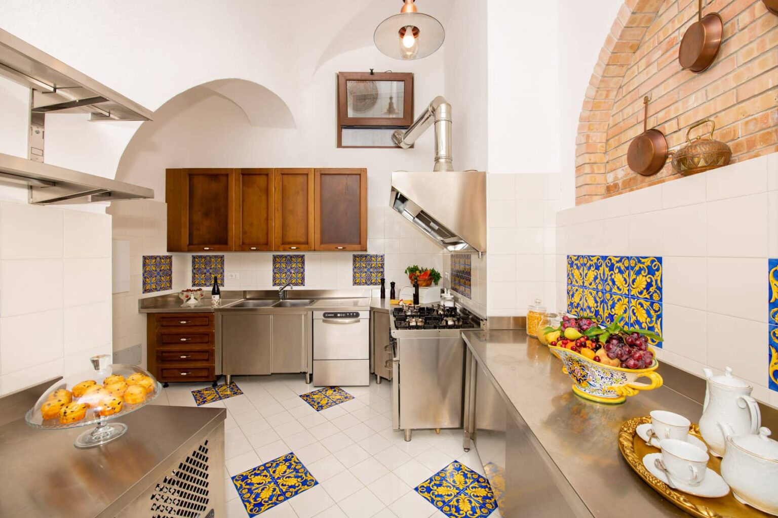 Modern kitchen interior featuring stainless steel appliances, wooden cabinetry, and colorful tiled accents. The space includes a fruit display and decorative elements, showcasing a blend of contemporary and traditional design. | Only on SopranoVillas
