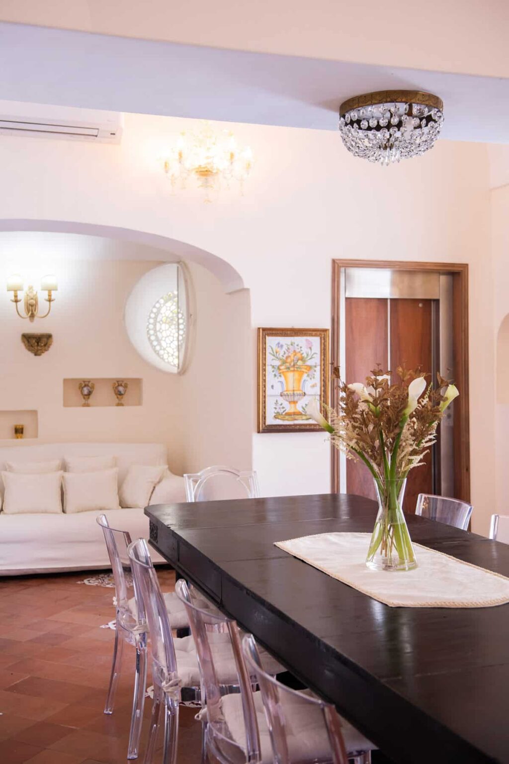 Modern interior design featuring a stylish dining area with a dark wooden table, clear chairs, and a decorative vase of dried flowers. The background includes a cozy living space with a white sofa, artistic wall decor, and elegant lighting fixtures. | Only on SopranoVillas