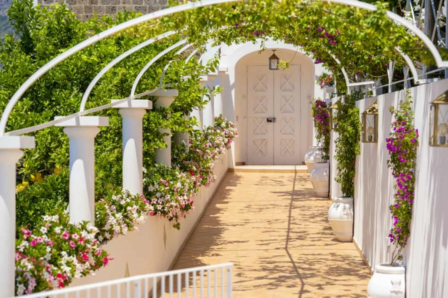 Scenic walkway adorned with vibrant flowers, white columns, and a charming door, surrounded by lush greenery and decorative lanterns, creating a picturesque outdoor setting. | Only on SopranoVillas