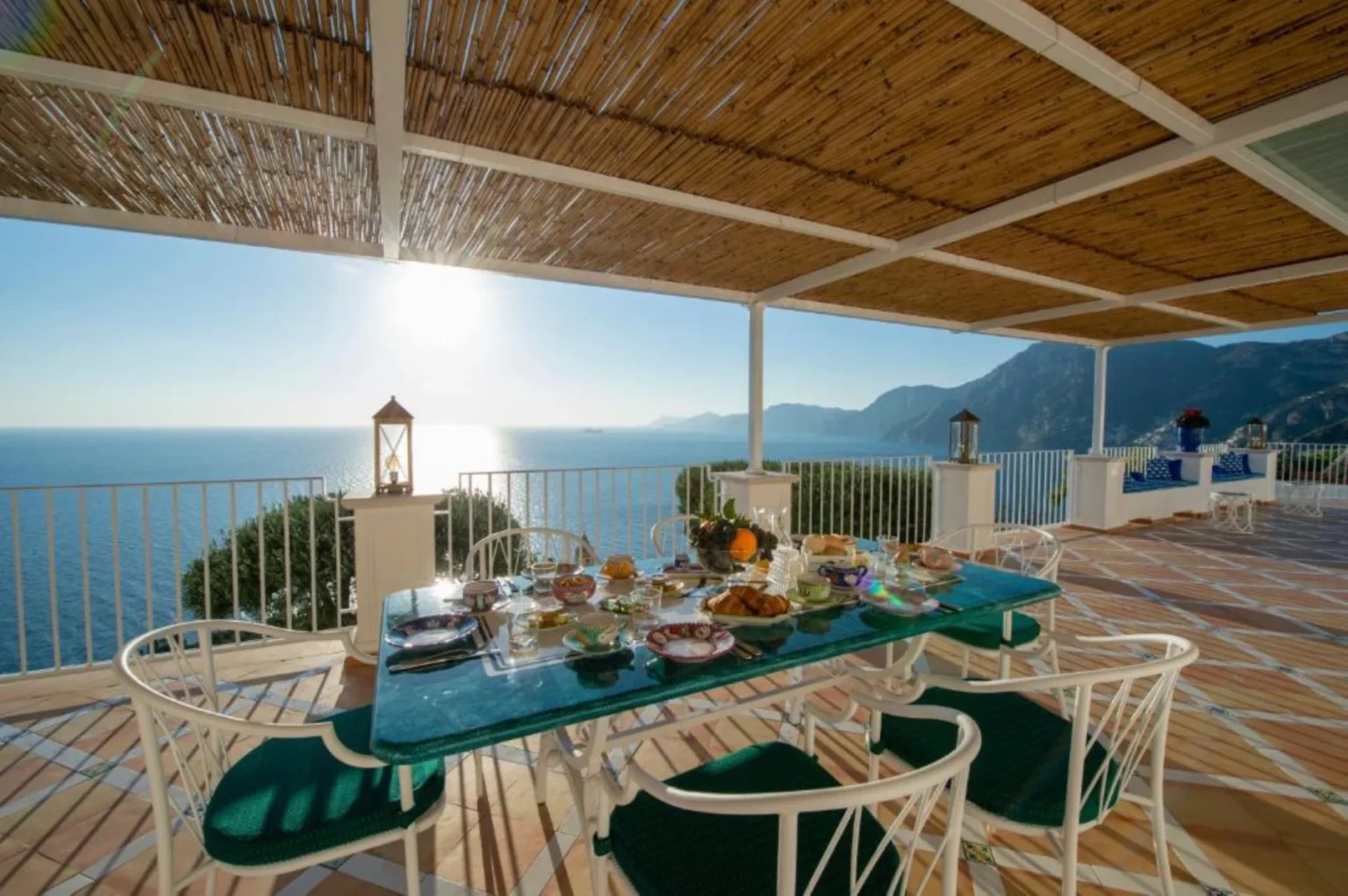 Stunning outdoor dining area with a table set for breakfast, overlooking a serene ocean view at sunrise. The scene features white wicker chairs, a green table, and a variety of delicious dishes, framed by a bamboo roof and coastal mountains in the background. Perfect for a tranquil getaway or vacation rental. | Only on SopranoVillas