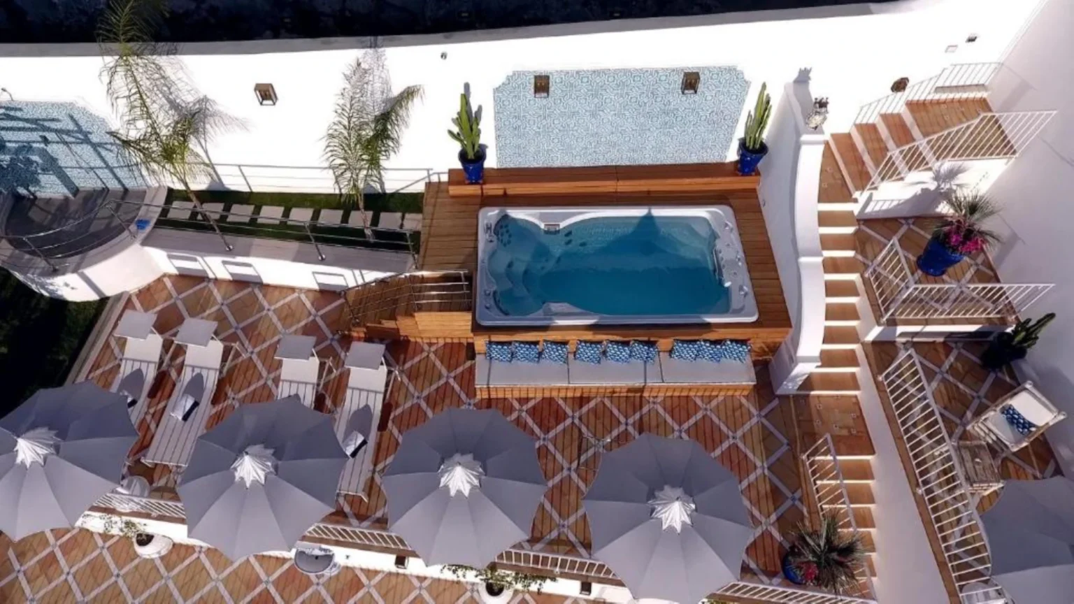 Aerial view of a luxurious outdoor space featuring a hot tub surrounded by wooden decking, lounge chairs, and sun umbrellas. The area is adorned with potted plants and a stylish tiled wall, creating a relaxing atmosphere for leisure and entertainment. | Only on SopranoVillas