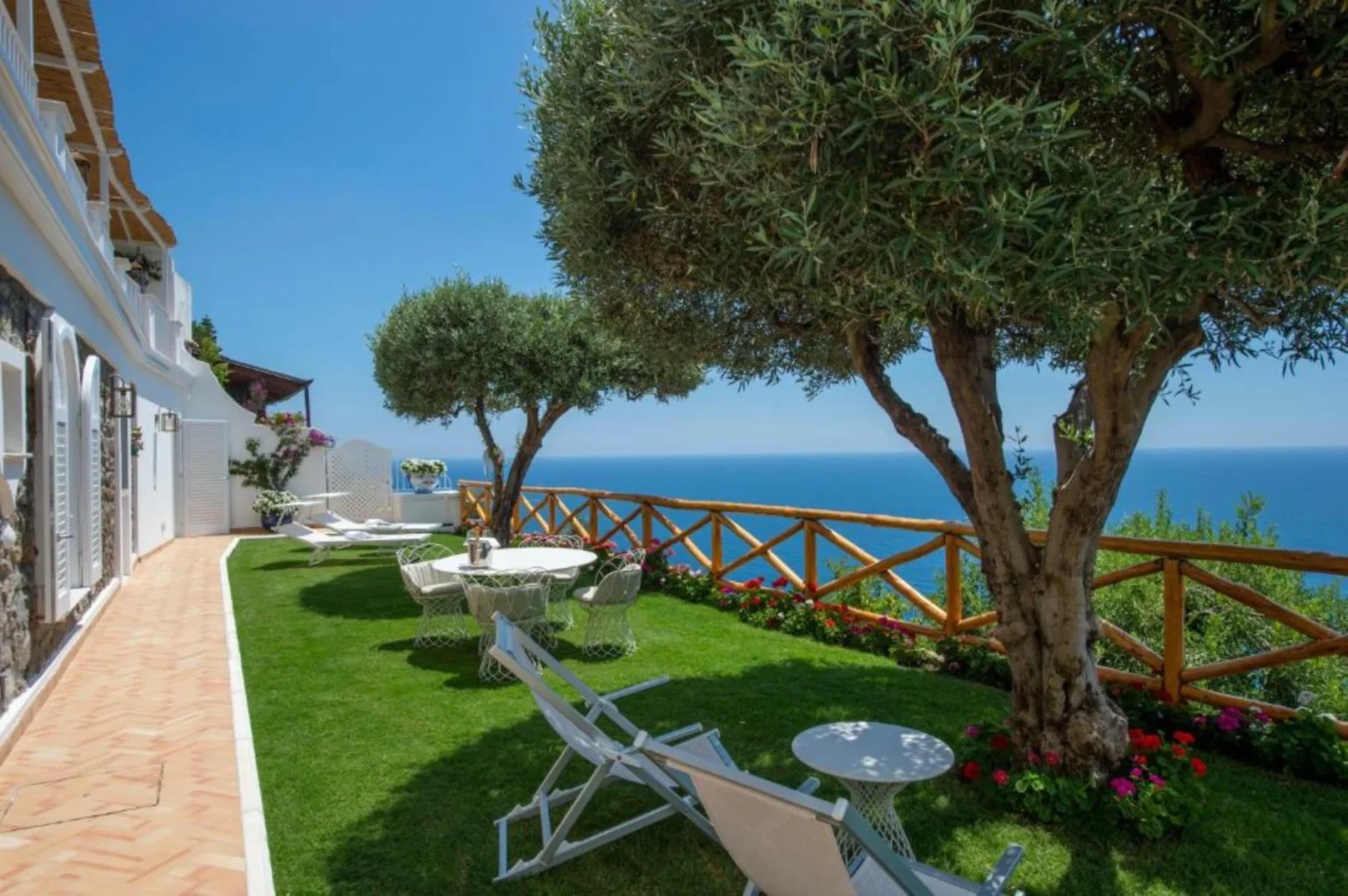 Scenic outdoor patio with olive trees, white lounge chairs, and a view of the ocean, surrounded by vibrant flowers and a wooden fence, ideal for relaxation and enjoying nature. | Only on SopranoVillas
