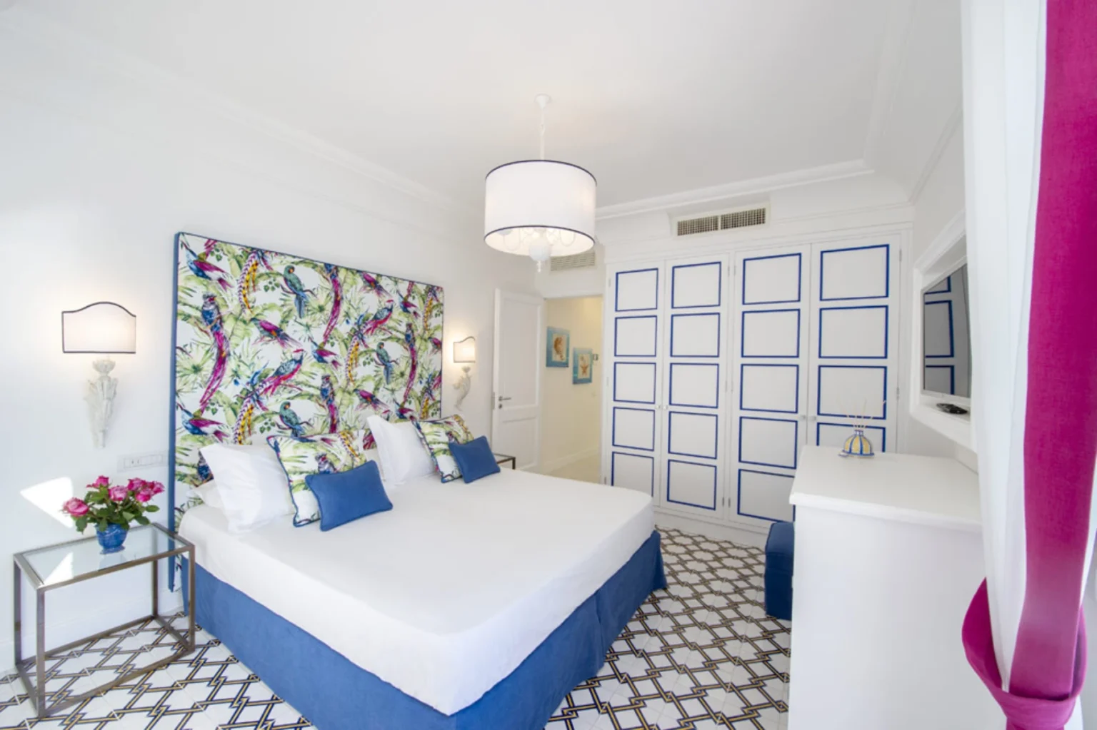 Bright and airy hotel room featuring a colorful floral headboard, white bedding with blue accents, a stylish floor with geometric patterns, and modern decor. Ideal for a relaxing stay, the room includes a bedside table with flowers and ample storage. | Only on SopranoVillas