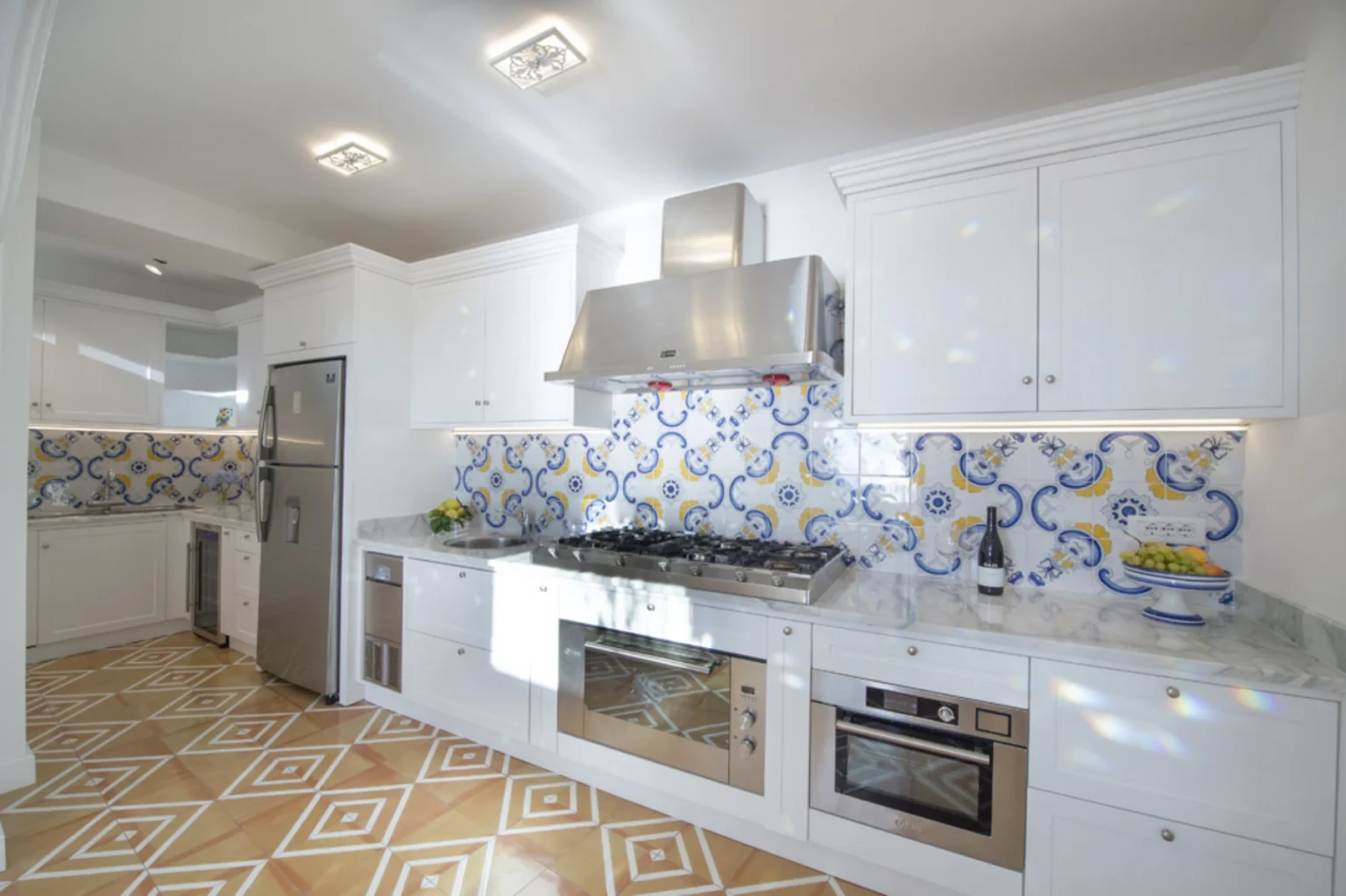 Modern kitchen featuring white cabinetry, stainless steel appliances, and vibrant blue and yellow patterned backsplash. The marble countertops complement the decorative tile flooring, creating an inviting culinary space. | Only on SopranoVillas