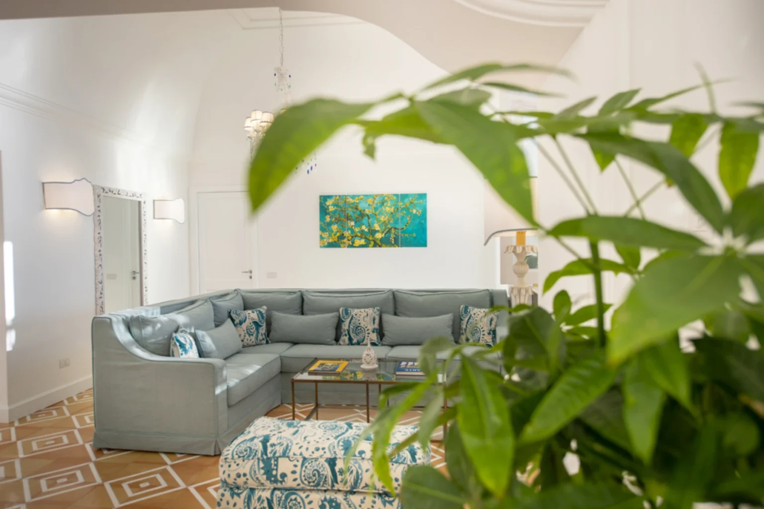 Bright and modern living room featuring a comfortable gray sectional sofa with decorative pillows, a glass coffee table, and a vibrant piece of artwork on the wall. Lush green plants add a touch of nature to the serene, well-lit space with stylish decor elements. | Only on SopranoVillas