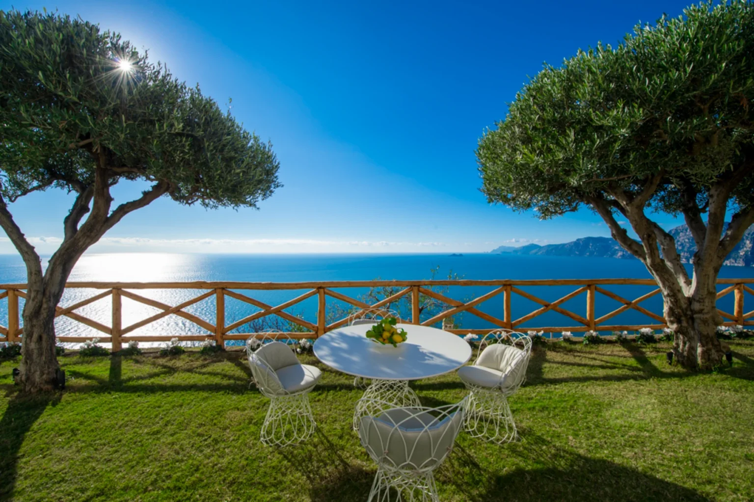 Scenic outdoor dining area with a round table and chairs overlooking a tranquil sea, framed by olive trees and a wooden fence, under a clear blue sky. Ideal for relaxation and enjoying coastal views. | Only on SopranoVillas
