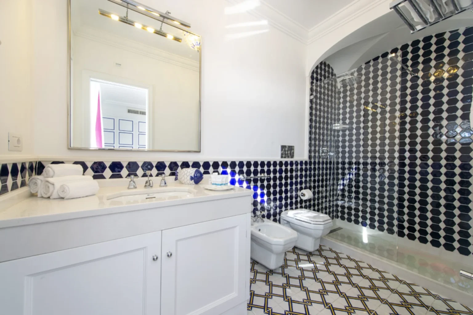 Modern bathroom featuring a stylish white vanity with a large mirror, neatly stacked towels, and elegant blue and white hexagonal tiles on the walls. The space includes a glass shower enclosure and contemporary fixtures, creating a chic and functional atmosphere. | Only on SopranoVillas