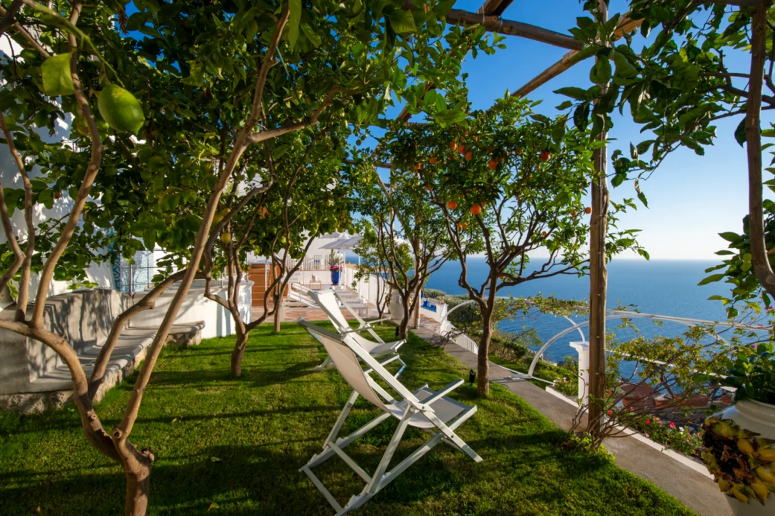 Lush garden with citrus trees and sun loungers overlooking the ocean, ideal for relaxation and enjoying scenic views in a coastal setting. | Only on SopranoVillas