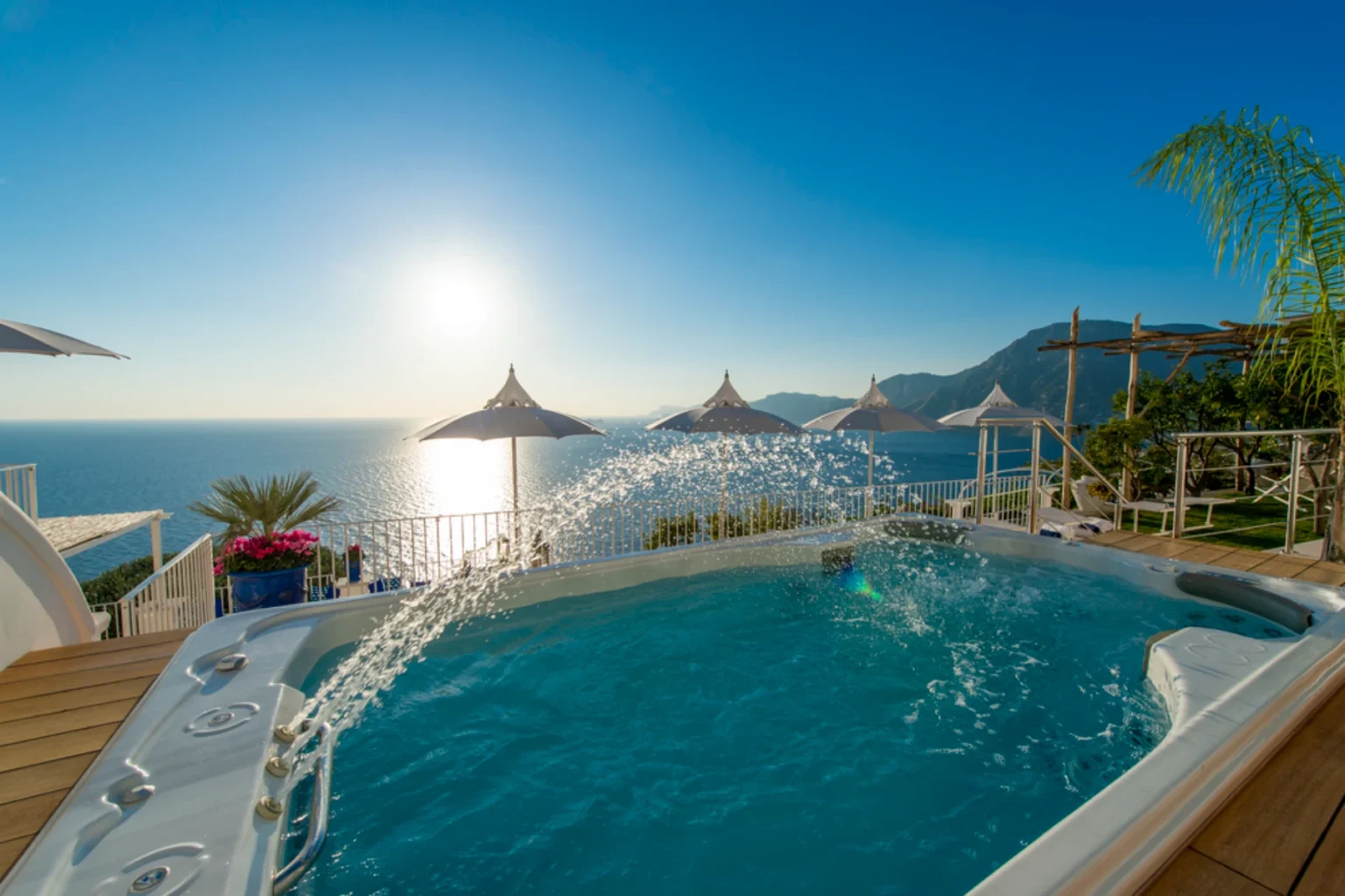 Scenic view of a hot tub overlooking the ocean at sunset, featuring white umbrellas and lush greenery, ideal for relaxation and luxury getaways. | Only on SopranoVillas
