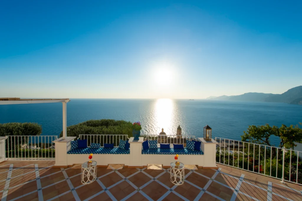 Scenic coastal view from a terrace overlooking the ocean at sunset, featuring blue seating, decorative lanterns, and vibrant greenery. Ideal for relaxation and enjoying breathtaking views. | Only on SopranoVillas