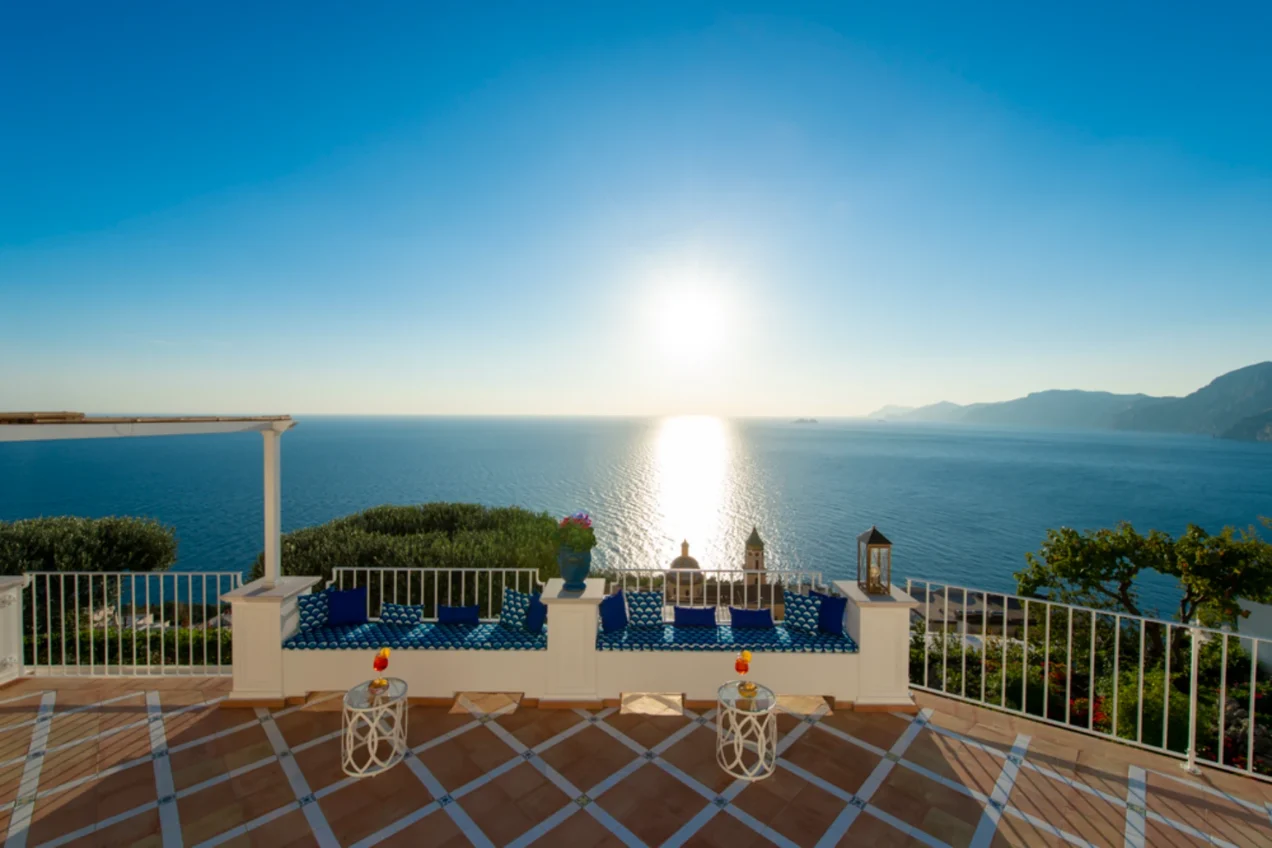 Scenic coastal view from a terrace overlooking the ocean at sunset, featuring blue seating, decorative lanterns, and vibrant greenery. Ideal for relaxation and enjoying breathtaking views. | Only on SopranoVillas
