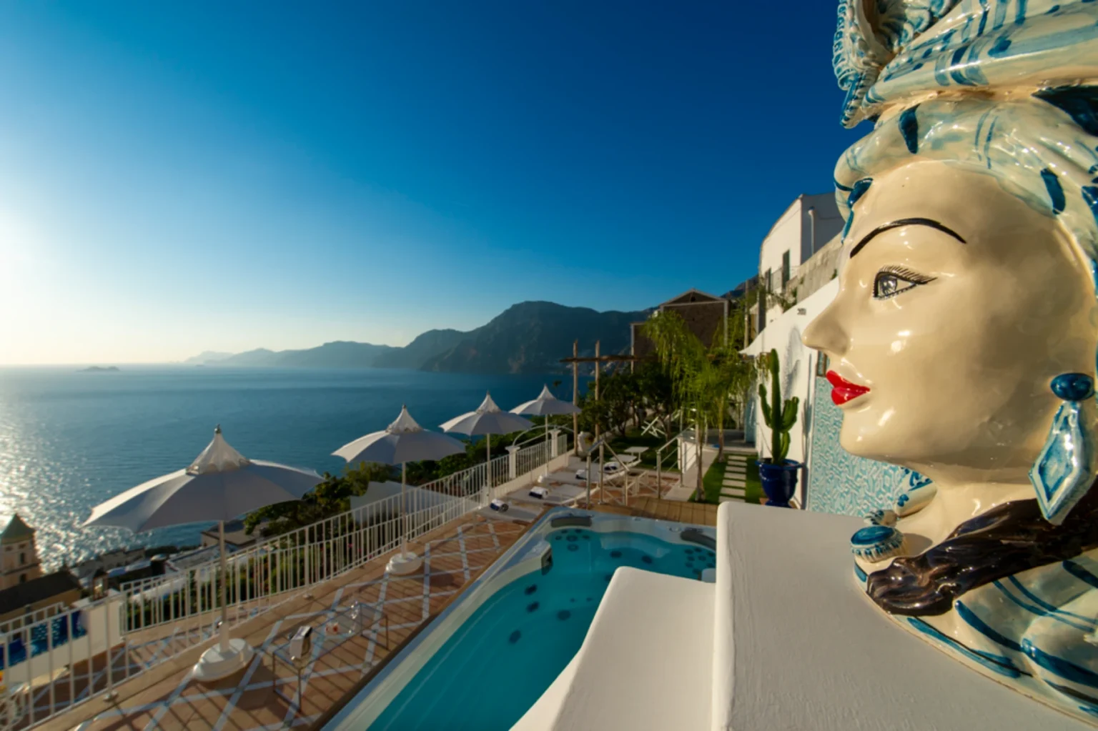 Stunning coastal view from a luxury villa featuring a decorative ceramic figure, a swimming pool, and white umbrellas against a backdrop of mountains and the sea, under a clear blue sky. | Only on SopranoVillas