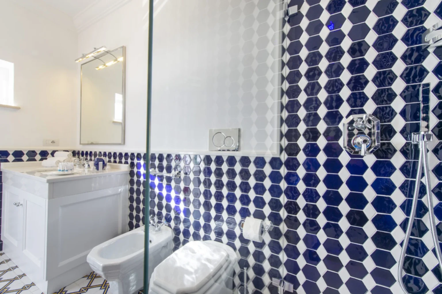 Modern bathroom featuring hexagonal blue and white tiles, a sleek white vanity with a mirror, a shower area with glass enclosure, and contemporary fixtures, creating a stylish and functional space. | Only on SopranoVillas